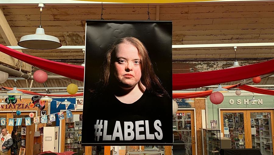 An exhibition photo of a young woman who has Down's Syndrome and is wearing a T-shirt with the word Labels on it. It hangs in a large hall