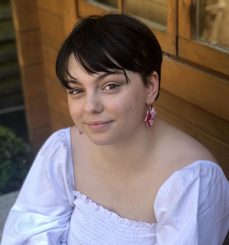New starter at Grapevine, Agatha Devlin. She has short dark hair and is wearing earrings and white top as she sits in the garden