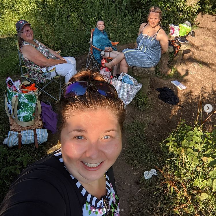 Four women sit in chairs together at the side of a river. One is at the front taking a selfie of them all