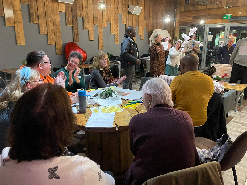 People sit and stand in a cafe either listening to or presenting ideas to those in the room.