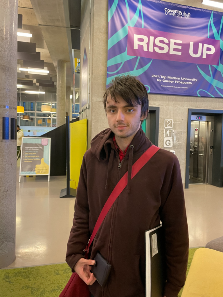 A young man wears a brown hoody with red crossbody bag and brown hair as he holds a notepad under his arm and stands inside Coventry University students union.
