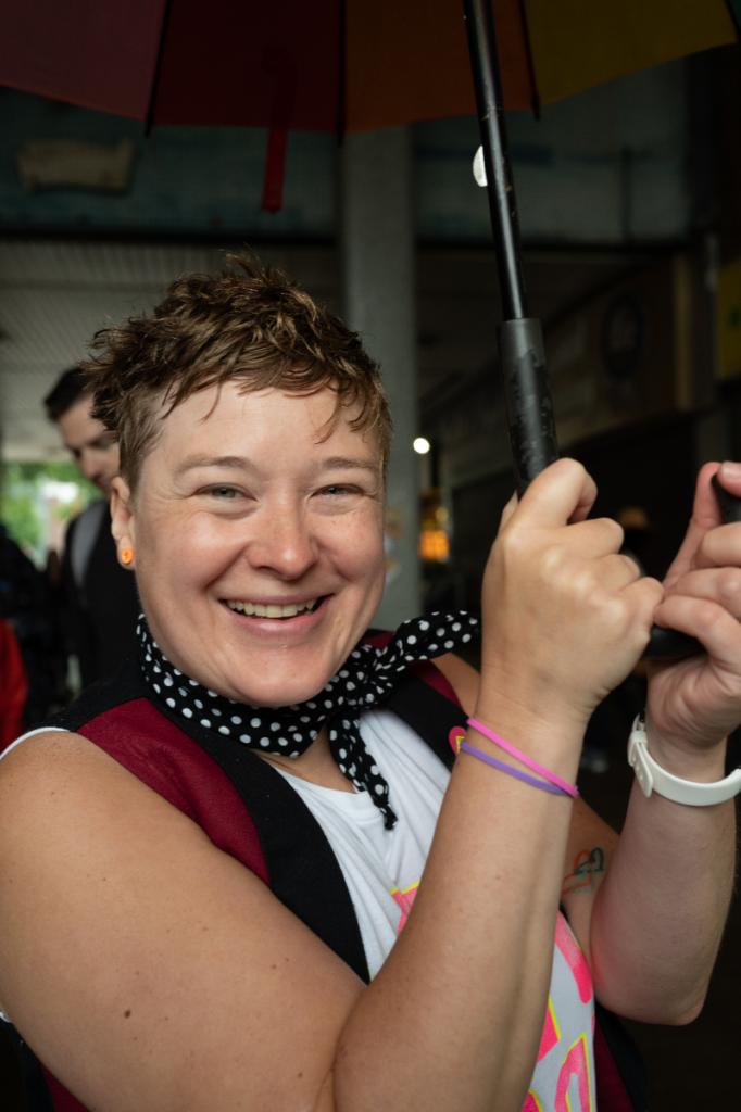 A white woman with short blonde hair wearing coloured bracelets and smiling broadly while sheltering under an umbrella.