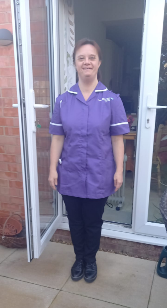 Joanne is seen smiling in her purple health care assistant's uniform oiutside some white patio doors. She looks proud.