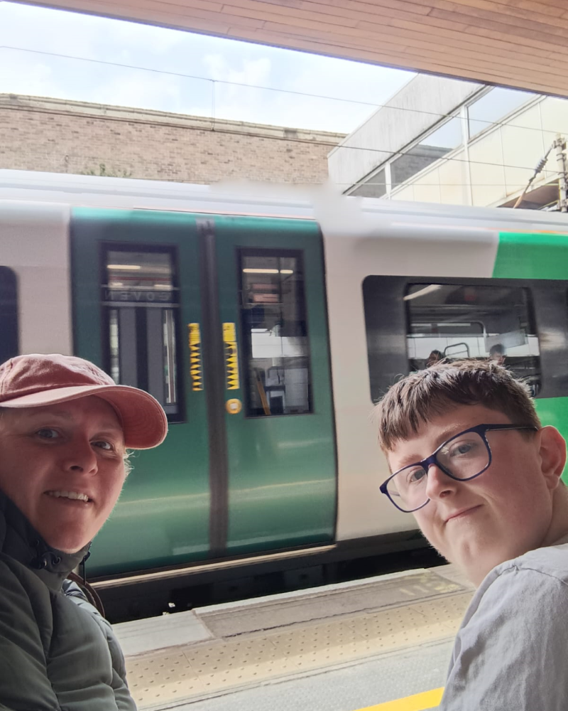 Michelle and Kirk sit either side of the camera lens with a train behind them that has stopped at the station. Michelle and Kirk are both white. Michelle wear a coat and pink cap and Kirk wears glasses with his short brown hair.