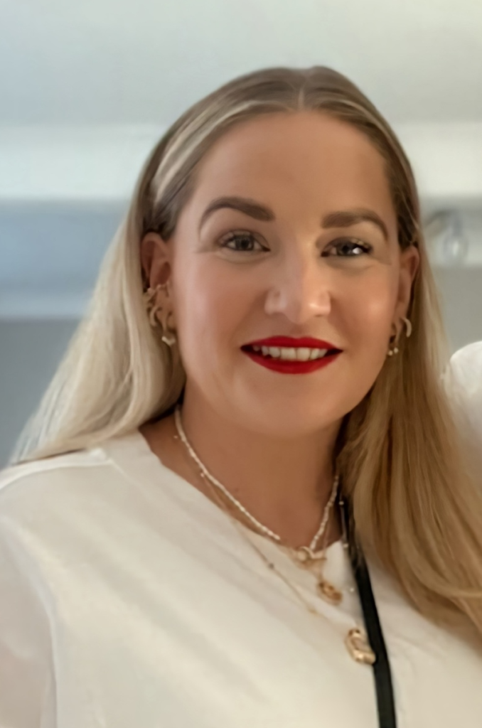 A white woman with long, straight blonde hair in her thirties wears red lipstick, lots of different gold earrings and a plain white t-shirt. She is smiling. This is Laura Fisher from Grapevine.