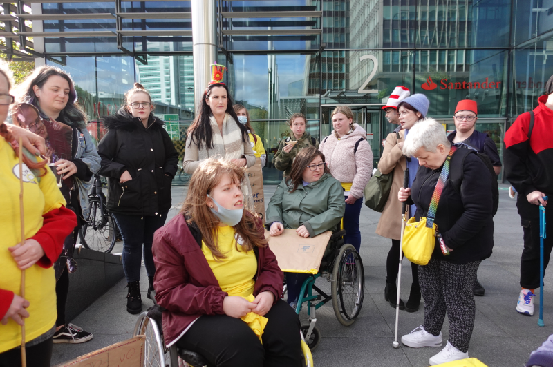 A large group of disabled activists sit and stand outside Facebook HQ in London armed with placards and tea ready to porotest about online disability hate