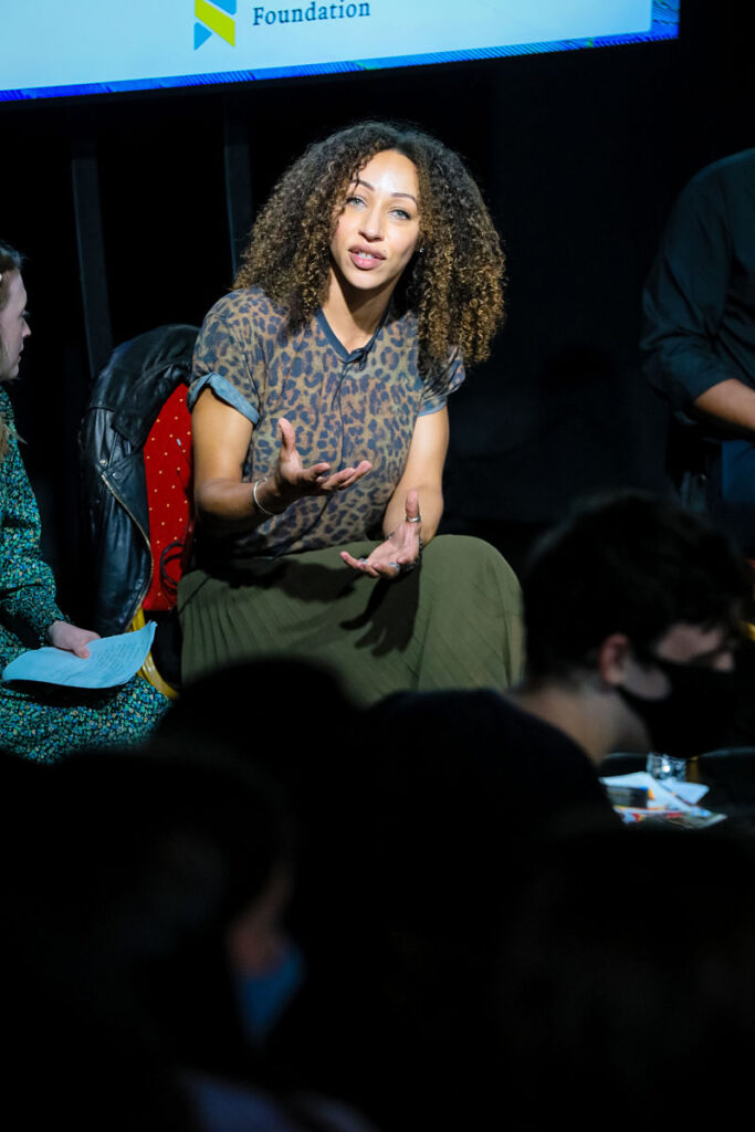 A woman with curly black hair and leopard print top sits on a stage panel speaking to the audience