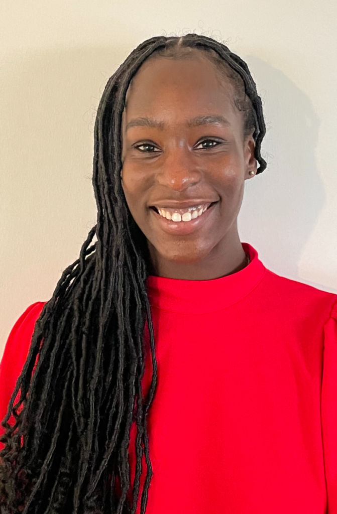 A young black woman wearing her briaded hair long over her shoulder and a red polo top, smiles for the camera against a plain background.