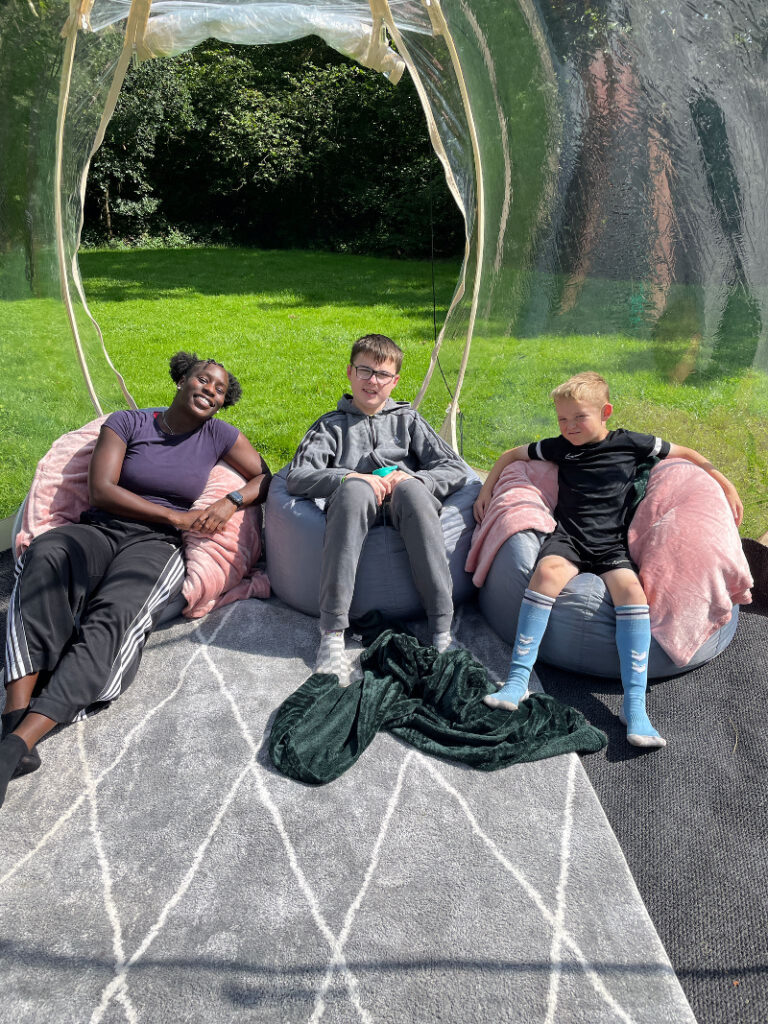 Three people, two young white boys and one young black woman, sit on bean bags inside an inflatable tent in a field. They are smiling in the sunshine.