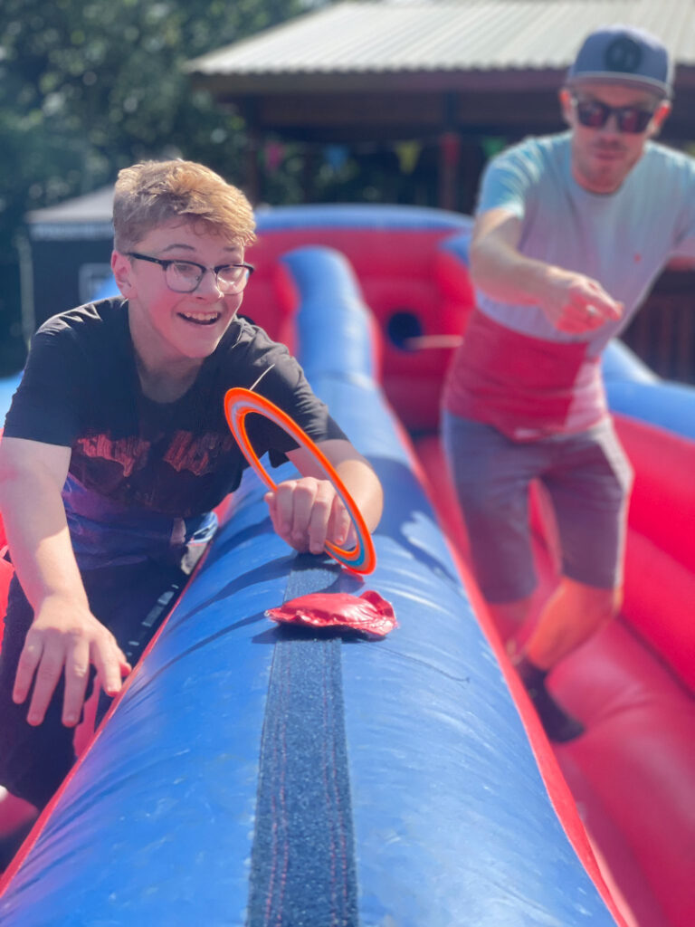 A young teenage boy with short auburn hair and glasses runs along a bungee run to grab a ring before the male project worker behind him.