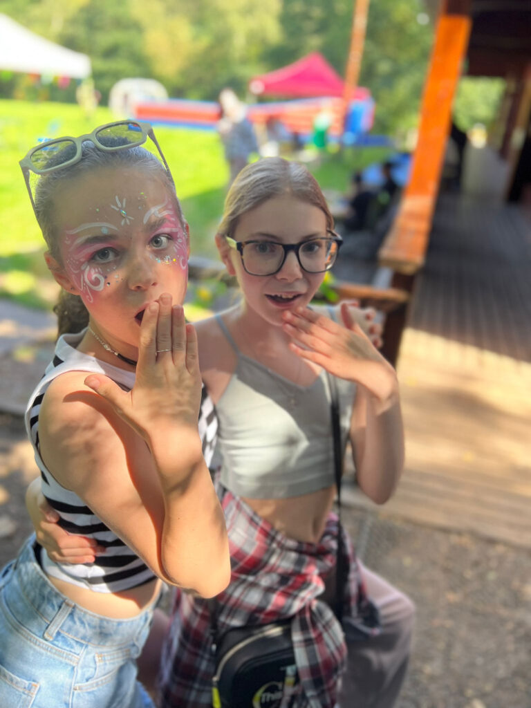 Two young white girls wearing glasses and summer vests pose for a photo outside the cabin. One has her face painted in pretty pastel colours. They both have long blonde hair tied back.