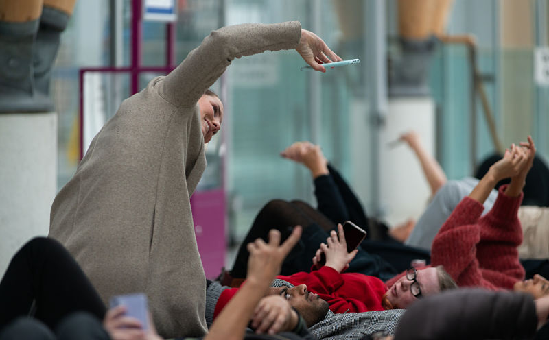 A woman in a beige coat takes photos of people laying on the floor