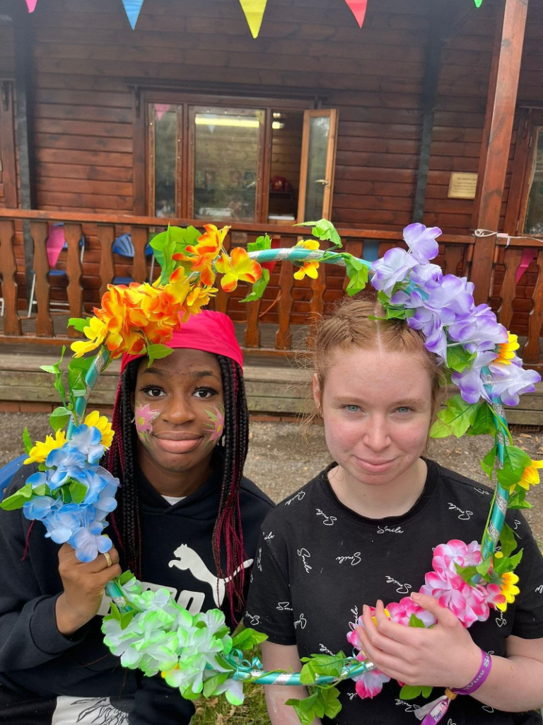 Two teenage firls, one black and one white, wear black tops and smile through a flower garland outside a log cabin at Teenvine's 2022 summer camp out.