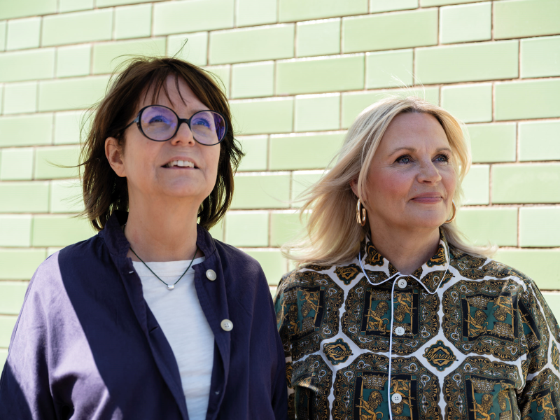 Two women stand side by side looking towards a clear sky outside the green tiled walls of Grapevine. The brunette woman wearing glasses is Clare Wightman and the blonde woman wearing a patterned green dress is Mel Smith.