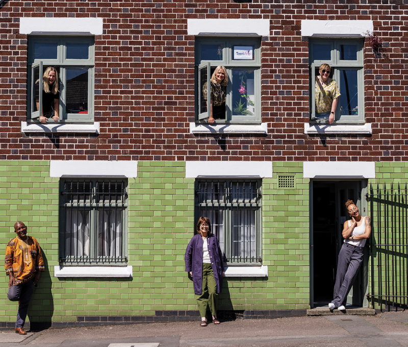 Photo of outside Grapevine's green tiled medieval building in Spon End, showing three women waving from open upstairs windows and two women and one man on the urban street below.