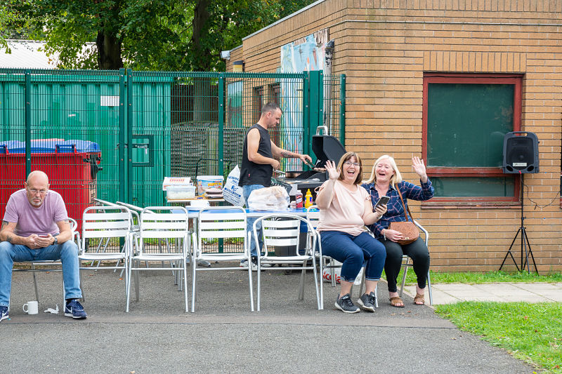 Two women sit on chairs while a man cooks food on the BBQ behind them. They are waving at the camera