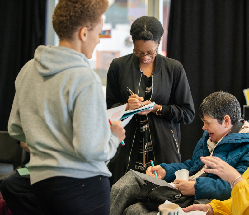 A young black woman takes notes as she speaks to an older disabled white woman who is sitting down and a younger woman of mixed ethnicity who is standing and wearing a light grey hoodie.