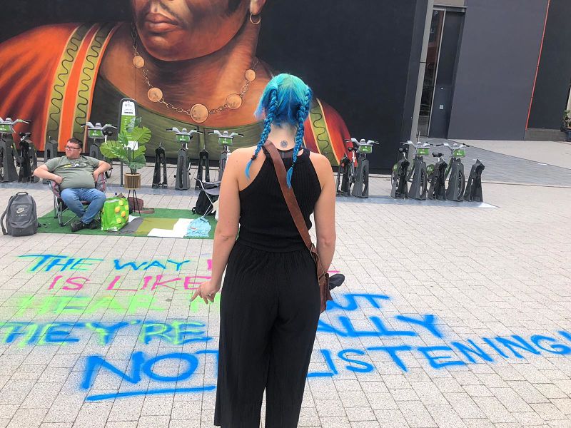A woman with blue hair in pigtails wearing black looks across a Grapevine parklet in Coventry city centre with faux grass, chairs and graffiti sprayed on the pavement