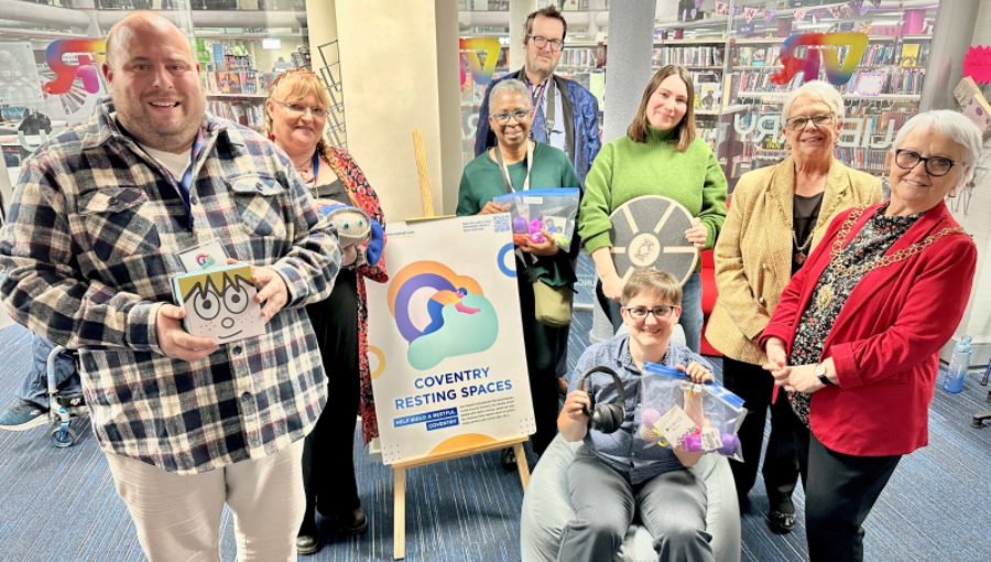 A group gathers inside the library for a publicity photo holding various sensory items available to loan and smiling for the camera. Credit: Coventry City Council.