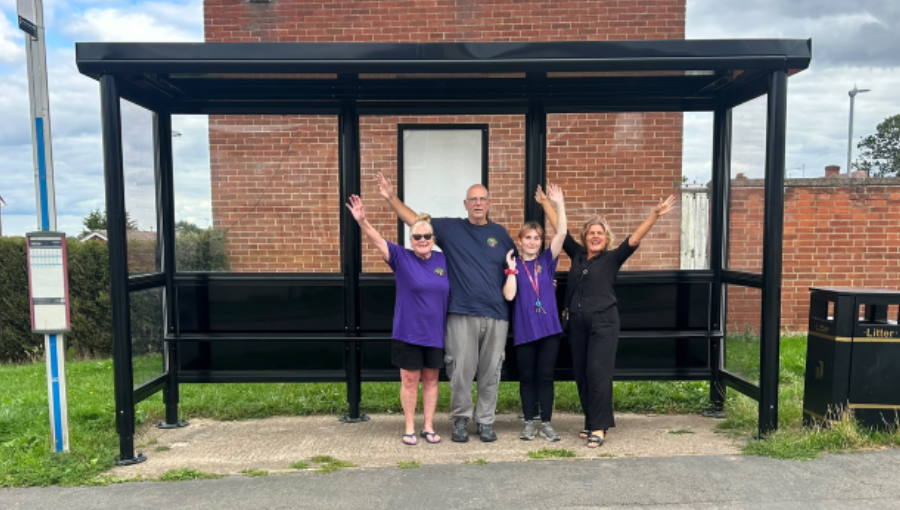 Three white women and one white man raise their arms in celebration under a new bus shelter in Nuneaton. They are some of the Friends of Grove Farm (left to right: Gwyn, Paul, Vicky and Grapevine community organiser Michelle).