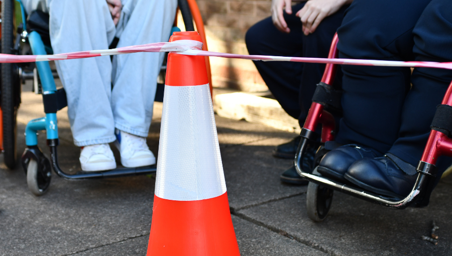 Photo of the bottom half of two people using wheelchairs in a street. A red and white traffic cone has tape attached either side to hold the disabled people back behind the line. Credit: Coventry Youth Activists.