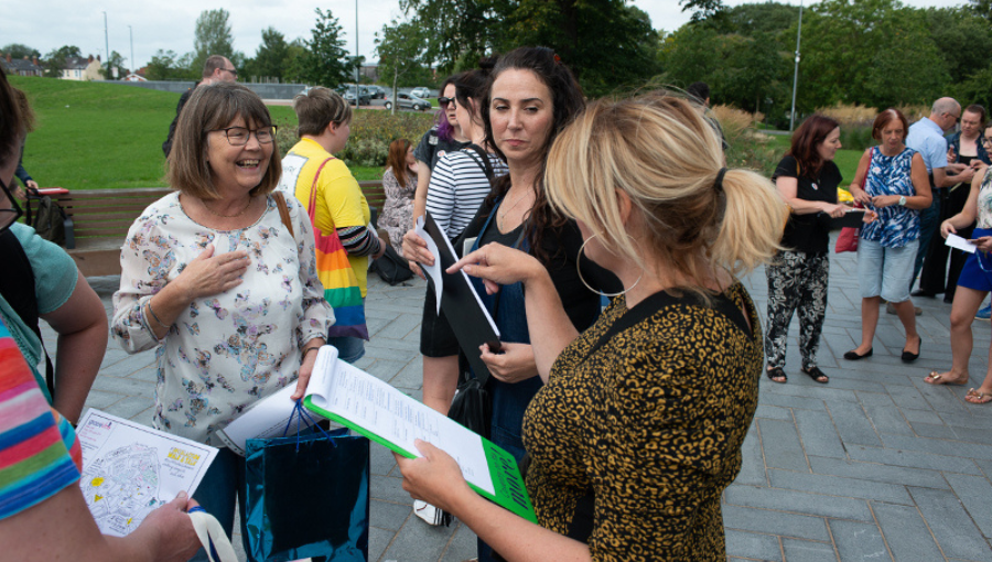 Three middle-aged white women in the foreground of an outdoor shot with a crowd behind. They are smiling and talking over a walk and talk plan.