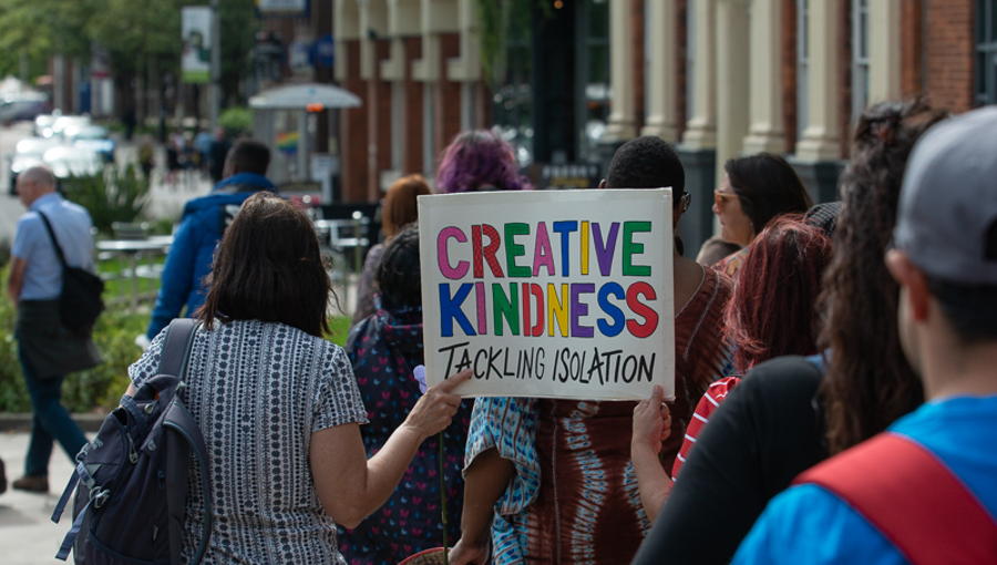 A group walks down a high street away from the camera holding a rainbow coloured placard that says, 'Creative Kindness tackling isolation'.