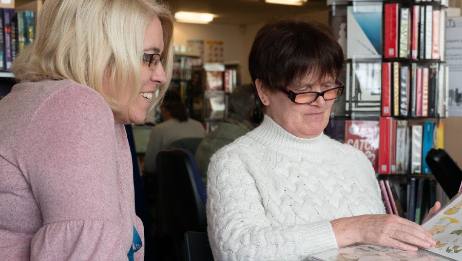 Two women in their forties, one brunette and the other blonde, sit at a table in a library looking at an art book together and smiling.