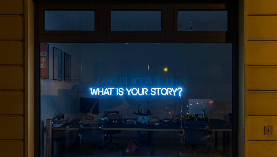 An office window at night with a neon sign hanging that reads: What is your story? Desks and computers are in darkness behind.