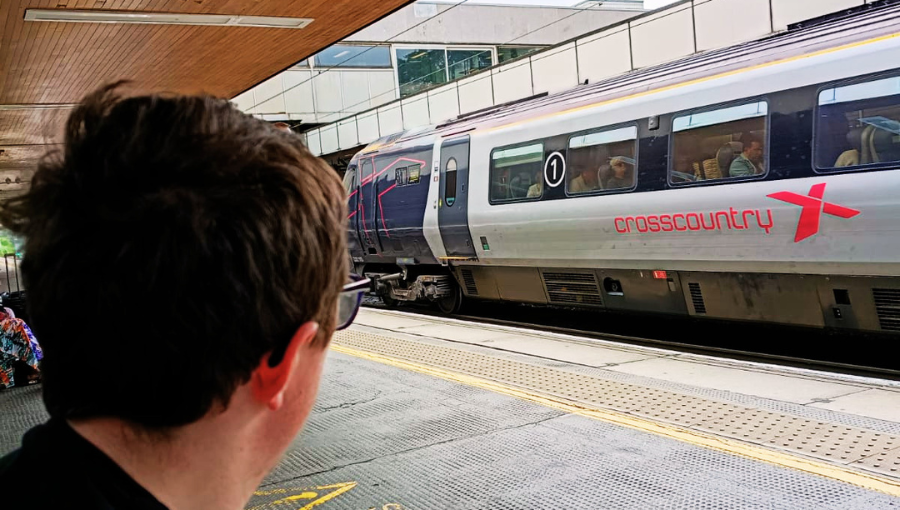 Photo shows the back of Kirk's head, with brown hair and glasses, as he watches a red and grey Cross Country train pass through the station past the platform where he is sitting.
