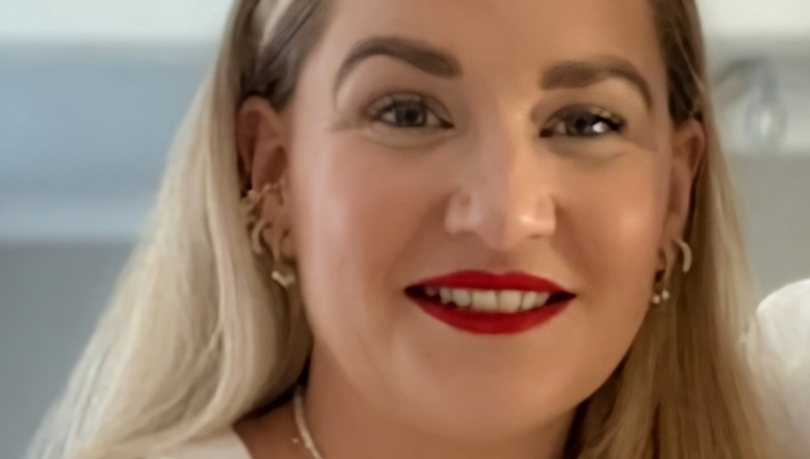 A white woman with long, straight blonde hair in her thirties wears red lipstick, lots of different gold earrings and a plain white t-shirt. She is smiling. This is Laura Fisher from Grapevine.