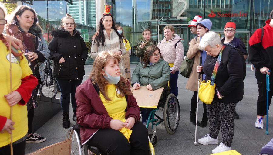 A large group of disabled activists sit and stand outside Facebook HQ in London armed with placards and tea ready to porotest about online disability hate