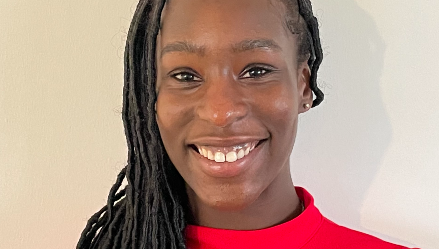 A young black woman wearing her briaded hair long over her shoulder and a red polo top, smiles for the camera against a plain background.