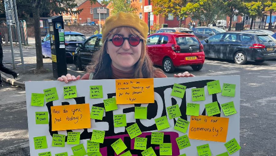 A young women with long hair wears a hat and sunglasses and holds a board covered in post-it note ideas from the community.