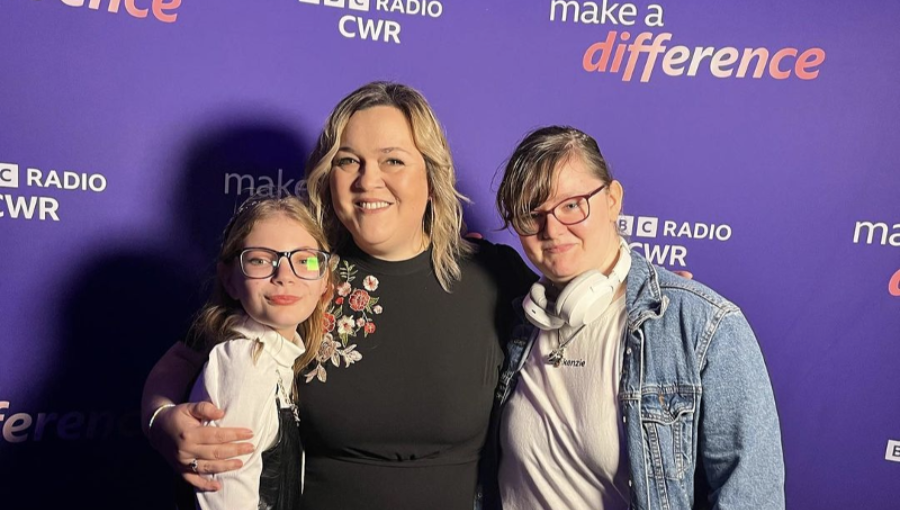 A woman with blonde hair wearing a black dress (Sarah) poses for a photo in front of a purple BBC CWR backdrop with young people Mayzee and Storm, two white teenage girls both wearing glasses.