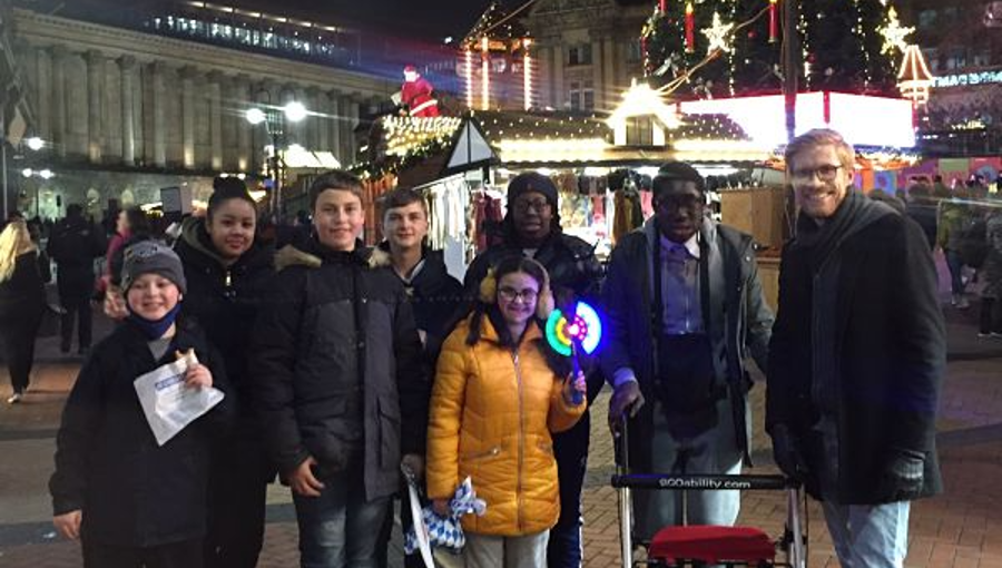 A group of teenagers with special educational needs and disabilities smile as they stand with project worker Paul in front of a huge Christmas tree at Birmingham's Frankfurt Market in December 2021.