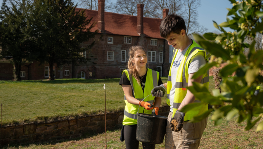 A young white woman and young white man wear yeloow tabards and rubber gloves as they share a black bucket while volunteering outdoors in a residential setting.