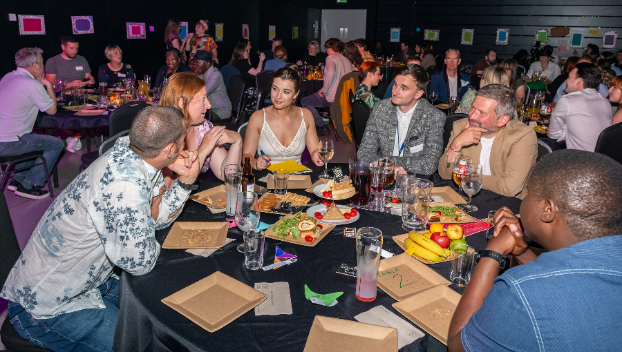 A large group of men and women sit in formal dress around an awards ceremony table with food and drinks laid out before them as they chat together.