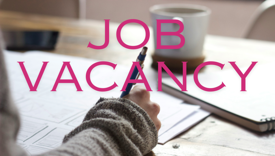 Someone's hand holds a pen to write on a notepad on a desk. In the background is a white coffee mug and words across the image say 'job vacancy' in bold pink letters.