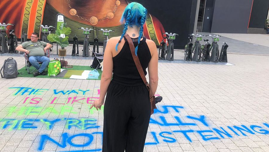 A woman with blue hair in pigtails wearing black looks across a Grapevine parklet in Coventry city centre with faux grass, chairs and graffiti sprayed on the pavement