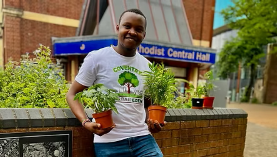 A young black man with short hair and a clean shaven face smiles as he holds potted plants next to a raised flower bed in Coventry city centre. He is Ade - a member of Coventry Urban Eden, whose branded white t-shirt he is wearing with blue jeans.