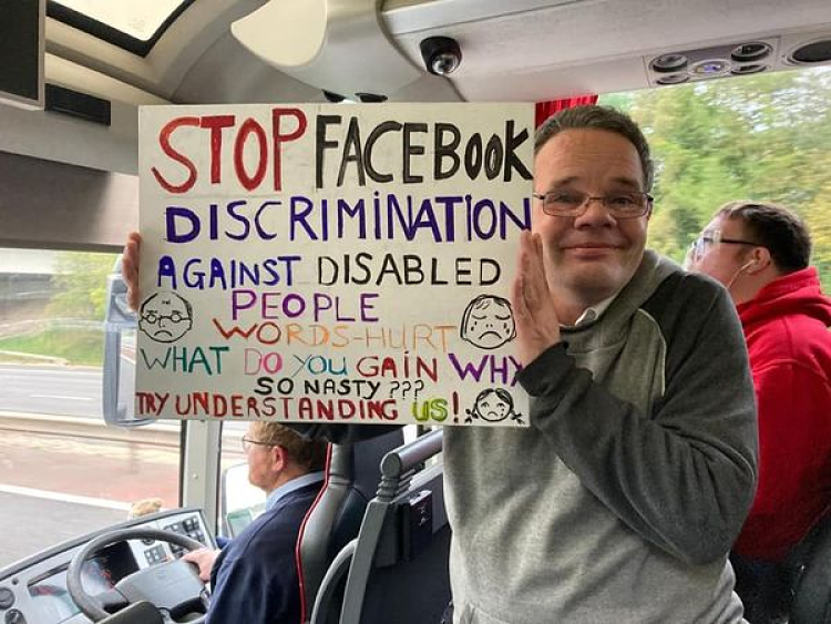 A CYA ally holds a cardboard placard about Facebook's disrimination of disabled people on the coach heading for their offices in London