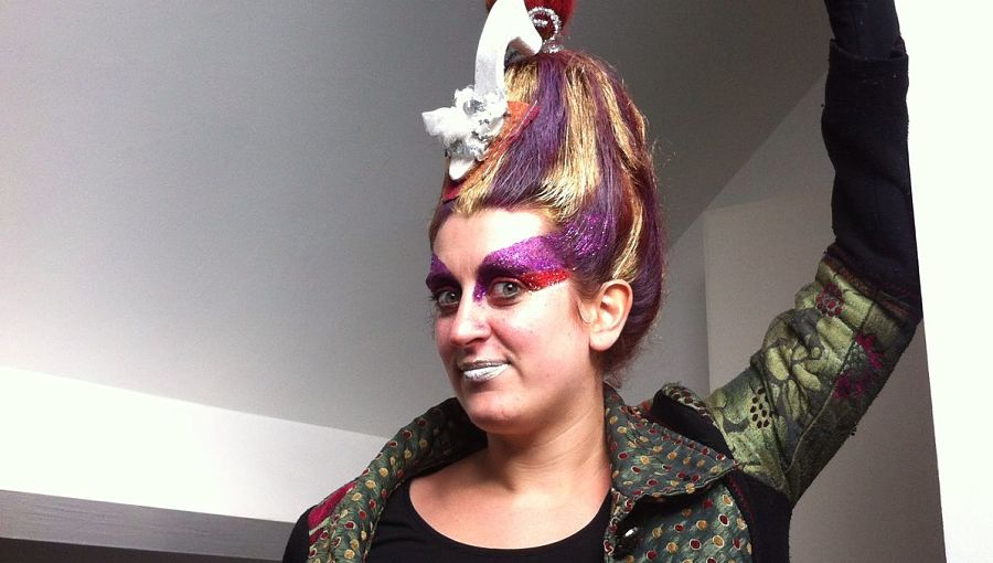 A woman with bright make up, colourful hair and costume dress smiles at the camera