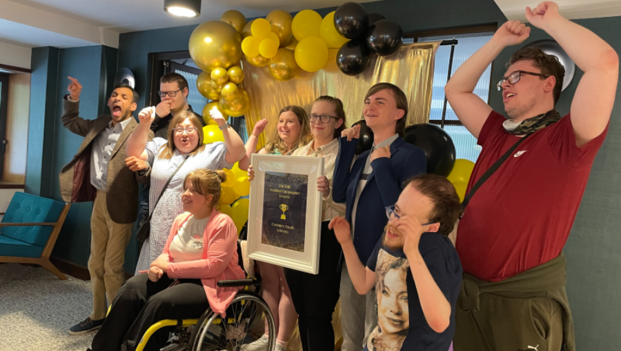 A group of disabled young people stand cheering and holding an award in front of yellow and black balloons. This is CYA celebrating after their 2022 SMK Campaigners national award win.