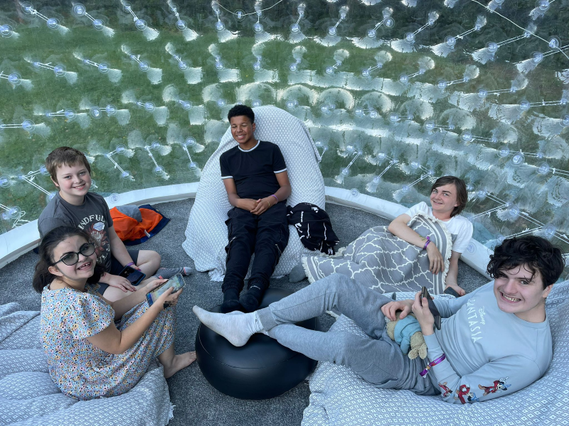 Five teenagers, boys and girls, sit chatting on beanbags inside a giant inflatable sensory bubble at Teenvine's summer camp out 2022.