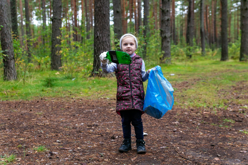 A young white girl with blonde hair is wrapped up for a litter pick in the woods. She holds a blue carrier bag and a piece of litter up for the camera.