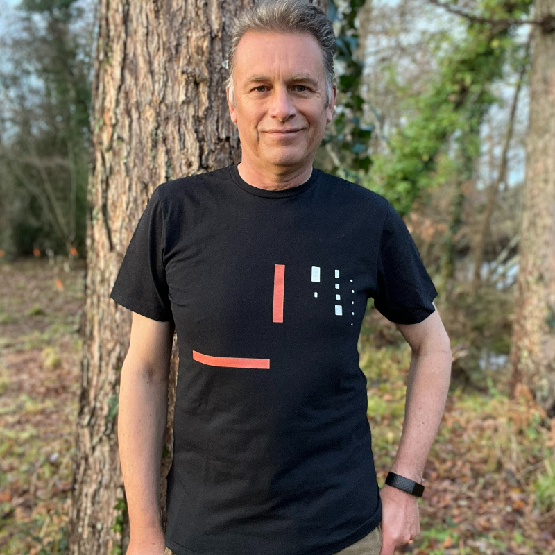 Chris Packham, a man in his fifties with blond grey hair, wears a black t-shirt and smiles in front of a tree in a woodland setting.