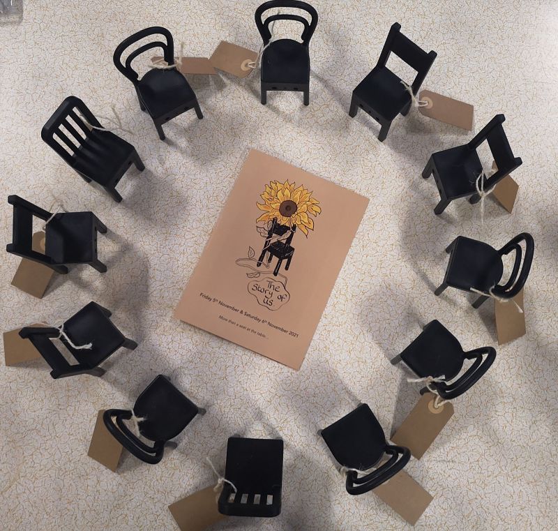 A set of black chairs of all different shapes form a circle around a card with a sunflower, a chair and the words Reform the Norm written on it