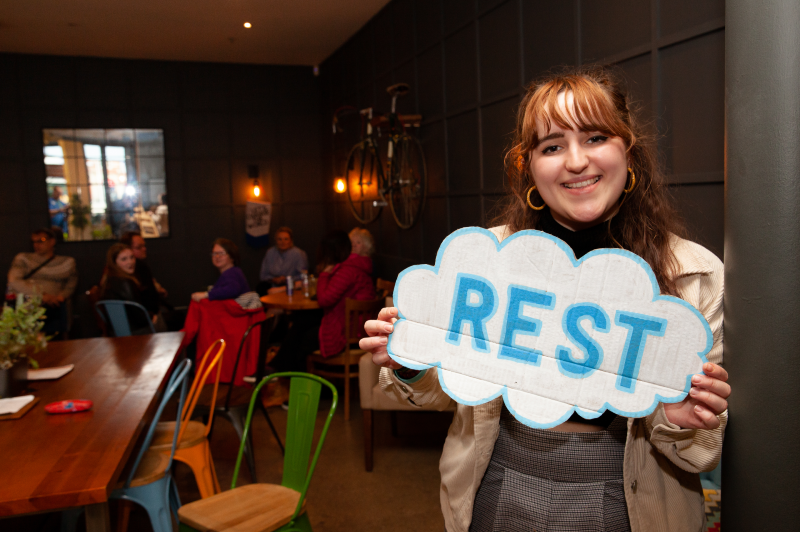 A young white woman with long brown hair and a fringe holds a handmade cloud sign with the word REST on it to the camera. Seated people behind her in the room turn to look at the camera too.