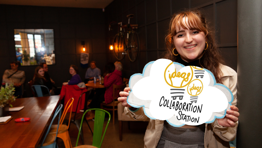 A young white woman with long brown hair and a fringe holds a handmade cloud sign with the Collaboration Station logo on it to the camera. Seated people behind her in the room turn to look at the camera too.